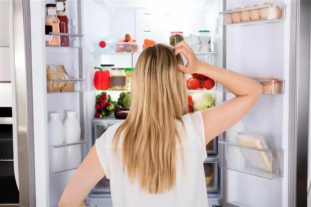 A view of a lady scratching her head standing in front of an opened fridge